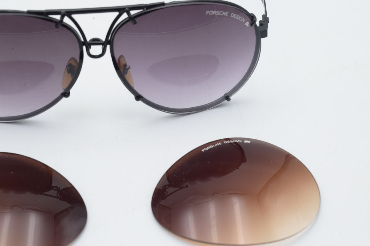 Black aviator sunglasses with dark gradient lenses, shown with two matching replacement lenses on a white background. The brand name 'PORSCHE DESIGN' is visible on the lenses.