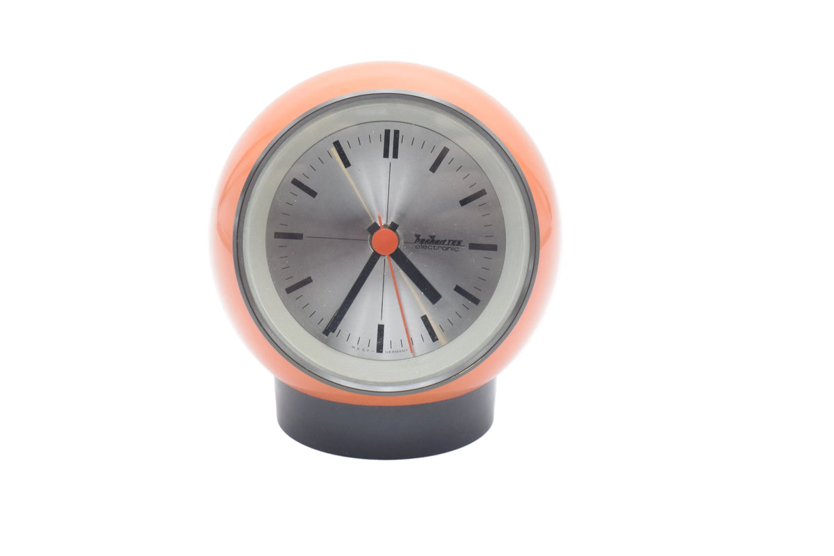 A retro-style spherical desk clock with a glossy orange shell, a silver-gray face, black hands, and a black base, shown on a white background.