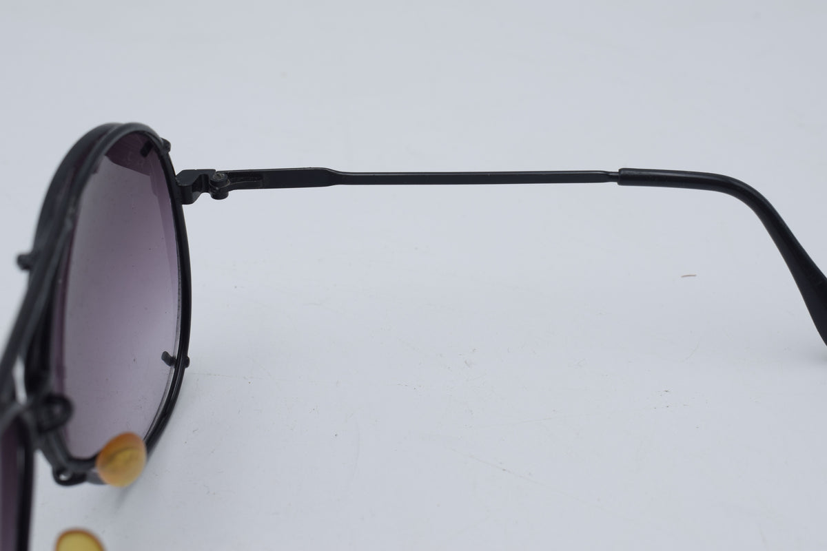 Close-up side view of a pair of sunglasses with a black metal frame and dark gradient lenses, resting on a plain light background.