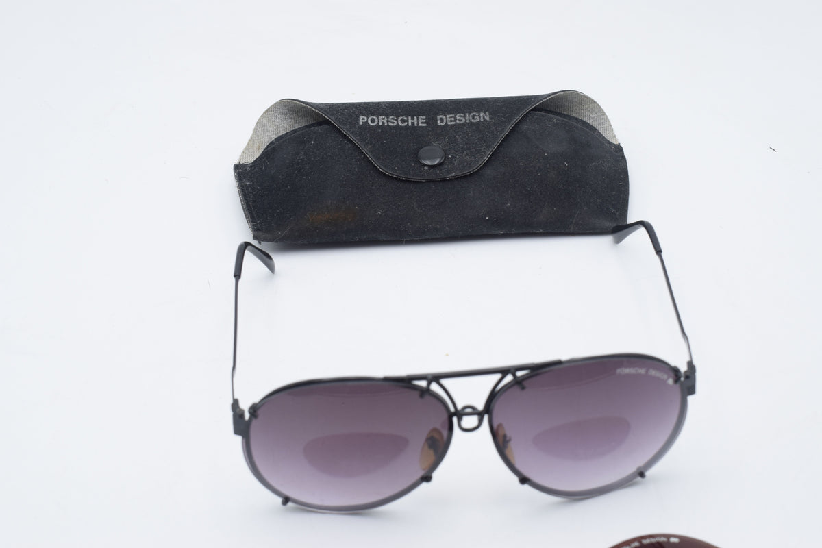 Porsche Design aviator sunglasses with dark gray frames and tinted lenses, displayed below their black suede case on a white background.