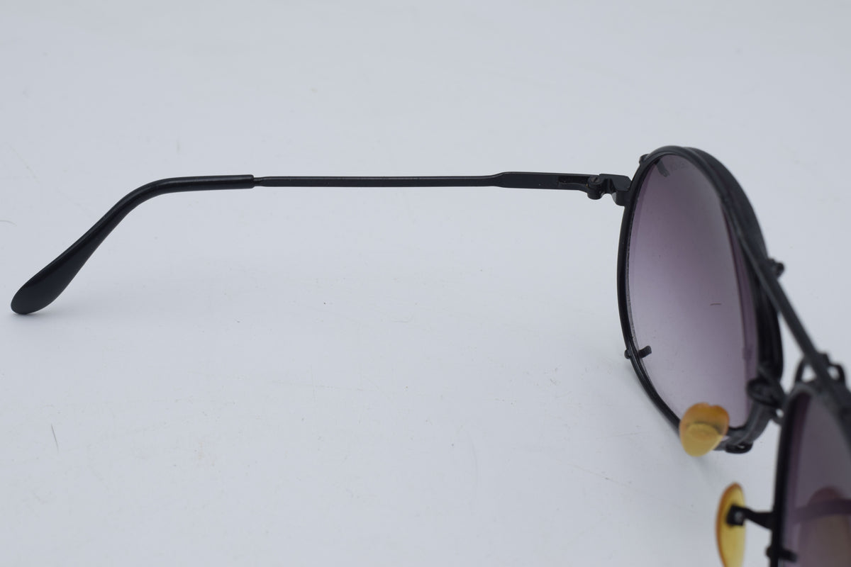 Close-up side view of a pair of sunglasses with a thin black metal frame, dark gradient lenses, and small gold-tone nose pads on a plain light background.