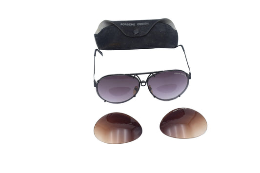 Porsche Design aviator sunglasses with dark gray frames and gradient purple-tinted lenses, shown with two matching replacement lenses and a black suede case on a white background.